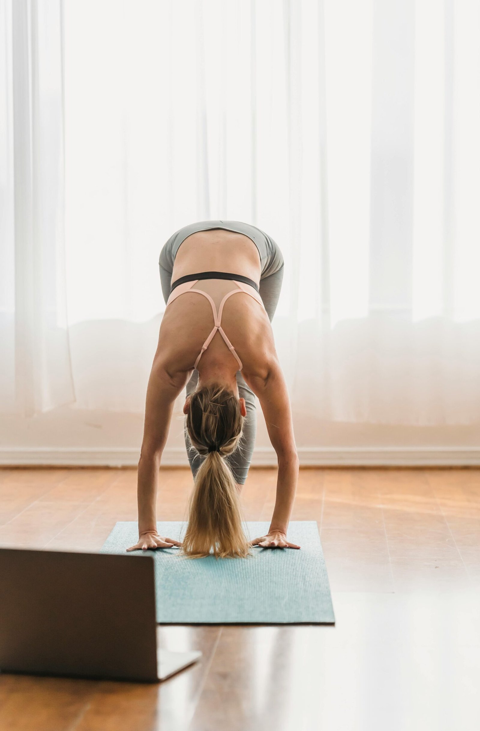 Woman doing a standing forward bend yoga pose at home with a laptop on the floor.